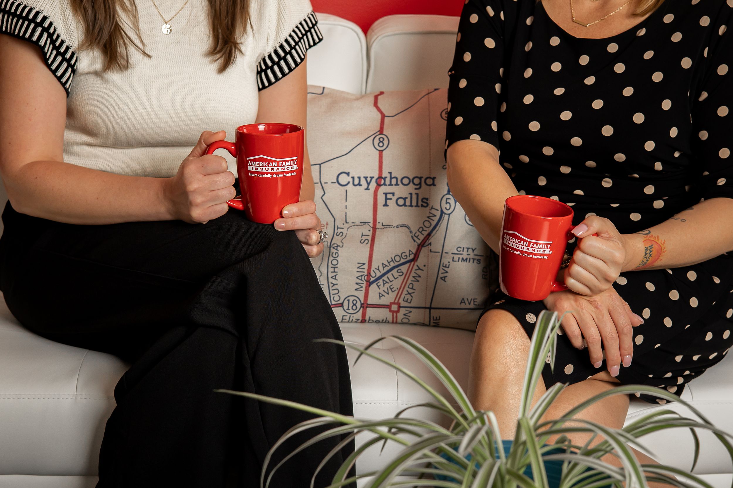 Two people sitting on a couch hold red coffee mugs with a plant and map visible in the scene.