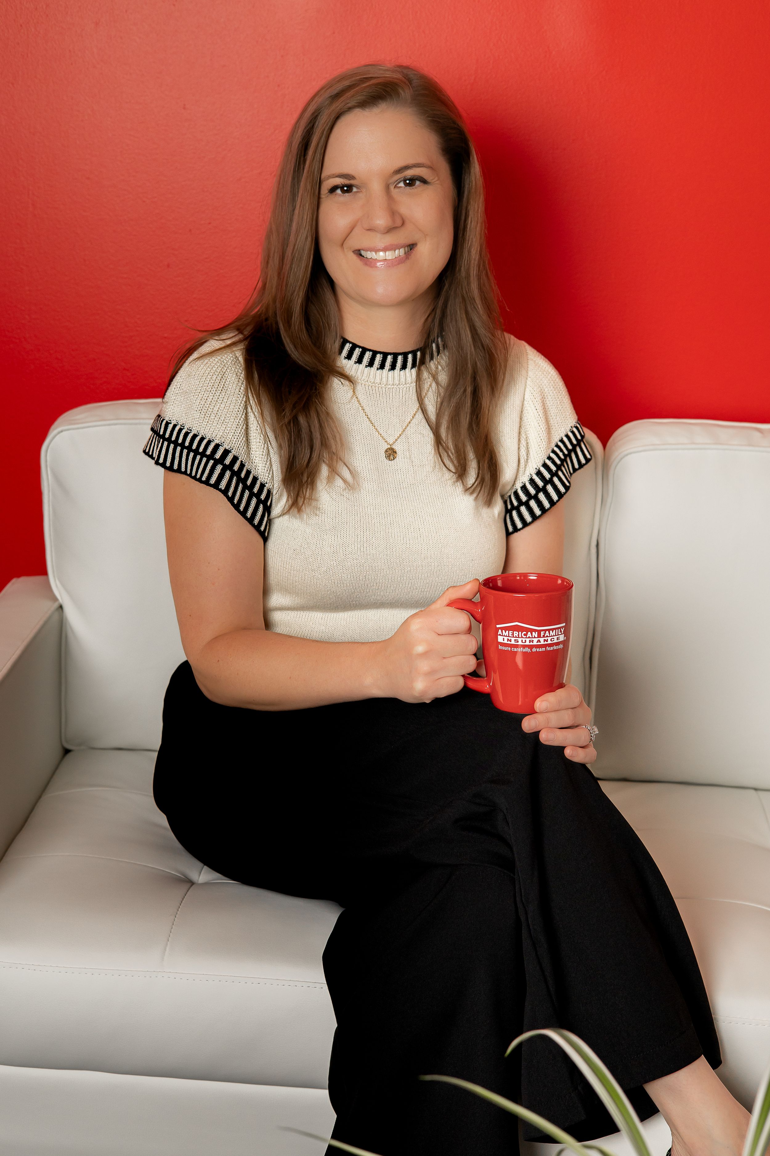 A person in a white top with black trim sits comfortably on a white couch against a red wall, holding a red mug.