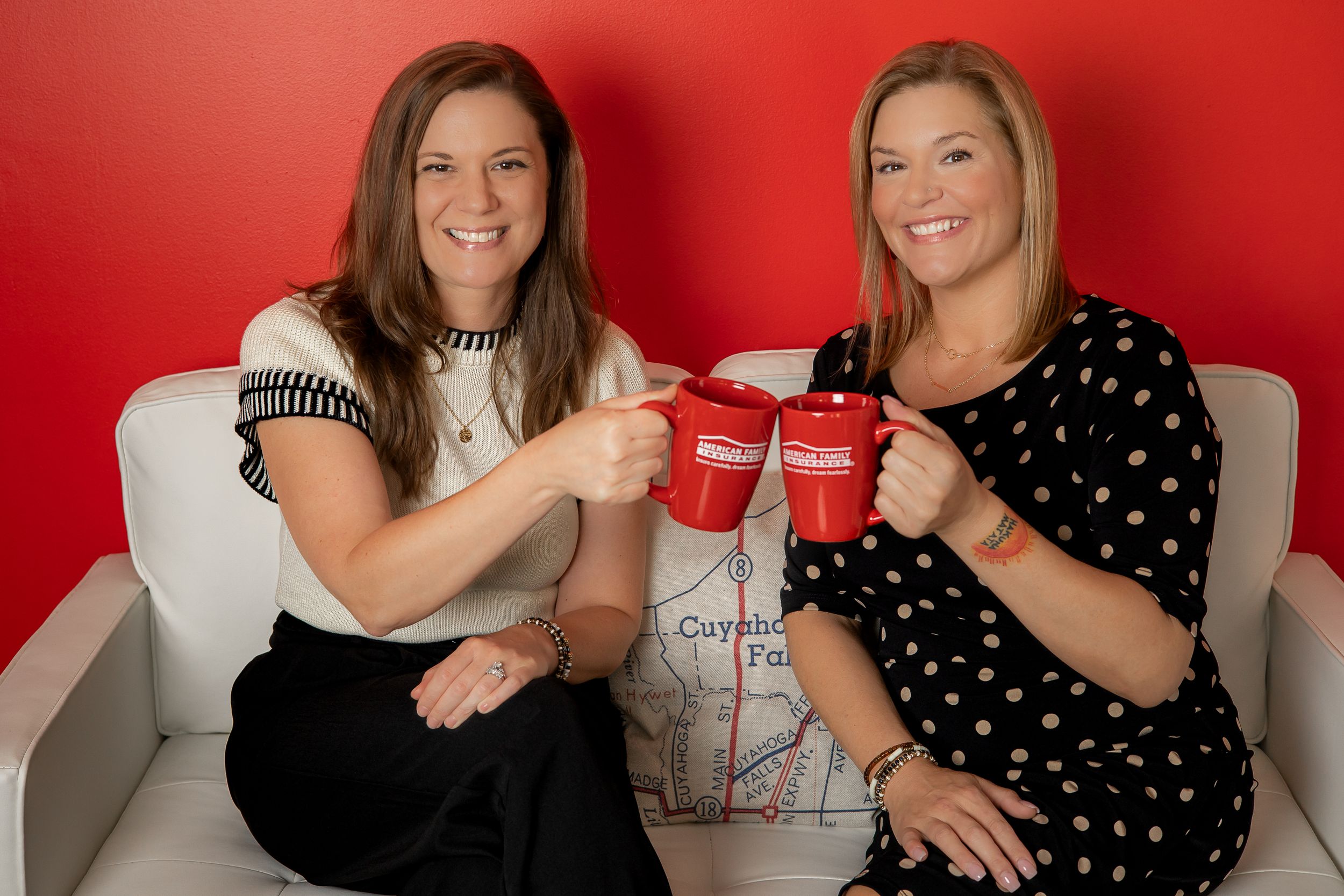 Two people sitting on a white couch toasting with red coffee mugs against a bright red background.