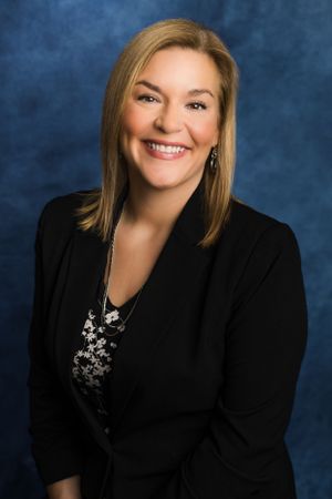 Professional headshot portrait with black blazer and floral top against blue background with warm smile and blonde hair.
