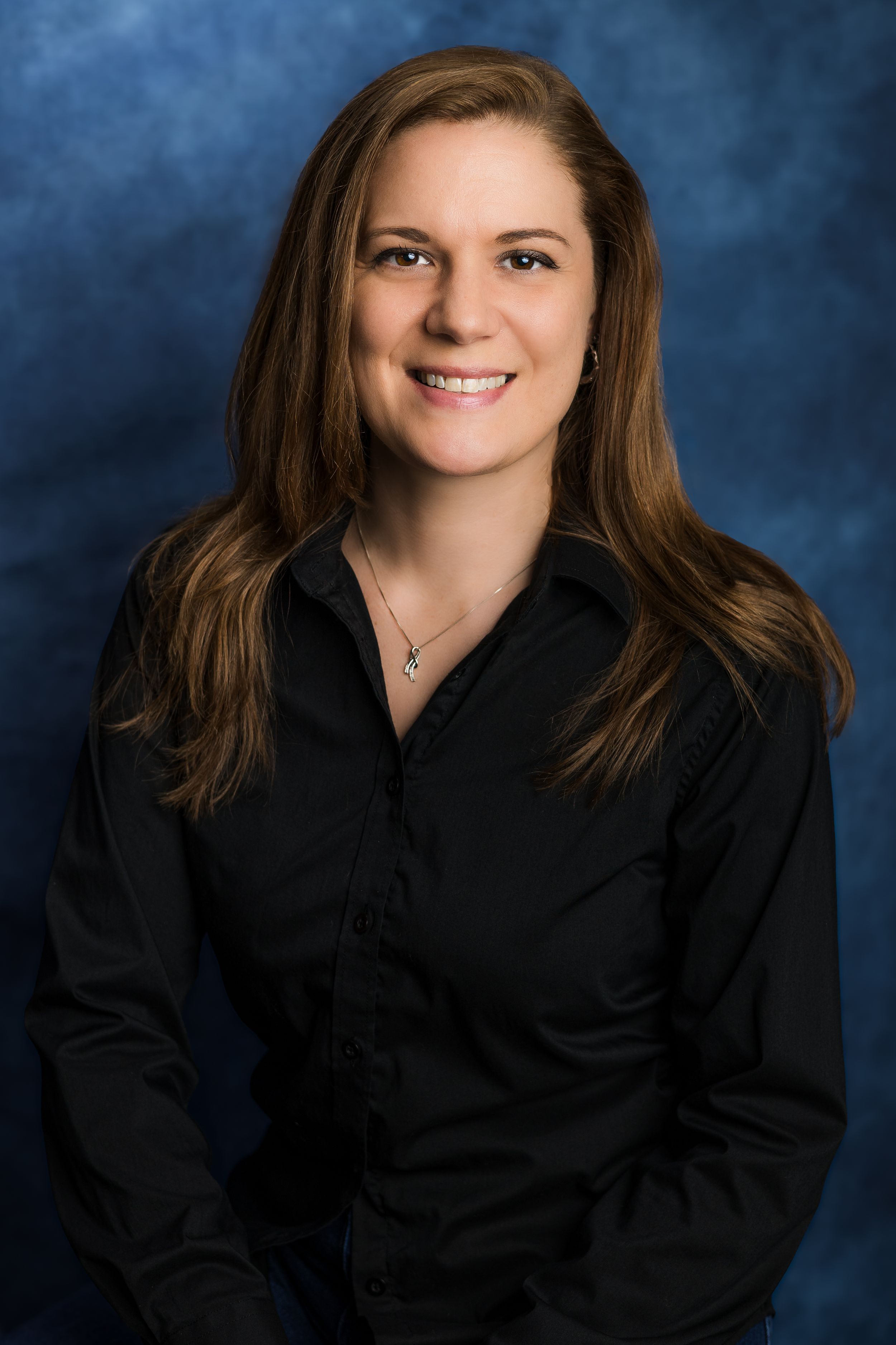 Professional headshot portrait against blue background showing person in black button-down shirt with long brown hair.