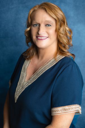 Professional headshot portrait of someone wearing a navy blue dress with decorative gold trim against a blue backdrop.