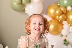 Smiling young girl with curly hair in lace dress surrounded by gold, white, and green balloons at a birthday celebration.