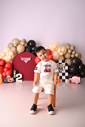 Toddler boy in white Cars outfit sits on chair with balloon garland and Route 66 racing themed birthday backdrop.