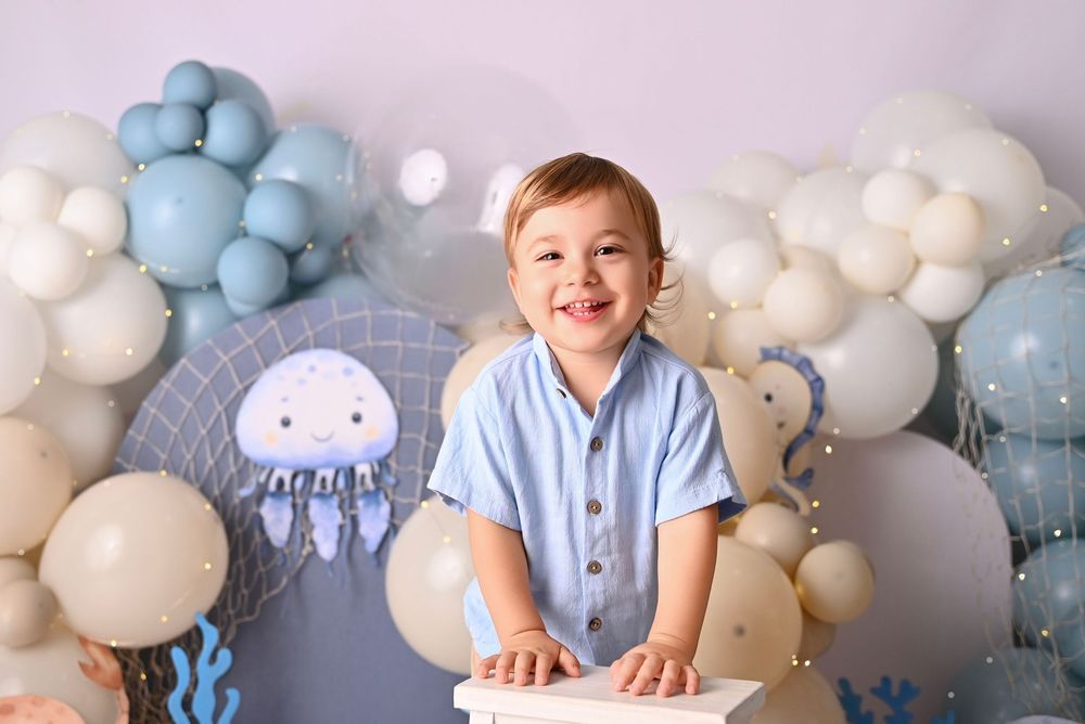 Smiling toddler boy in blue shirt at first birthday party with ocean-themed balloon backdrop featuring jellyfish decor.