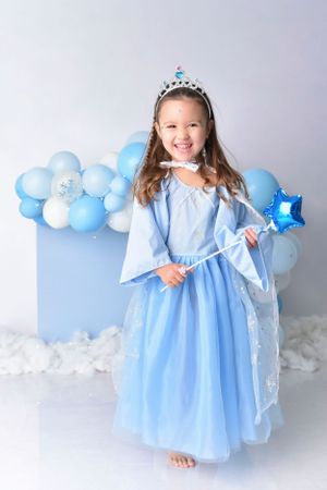 Young child in light blue fairy dress poses with matching balloons against white backdrop for magical photo shoot.