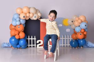 Birthday photo with balloon arch featuring child jumping from white chair.