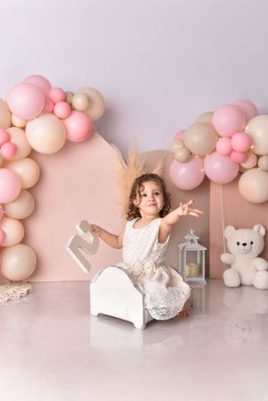 Girl in white dress poses on small bed with pink balloon arch and teddy bear decorations.