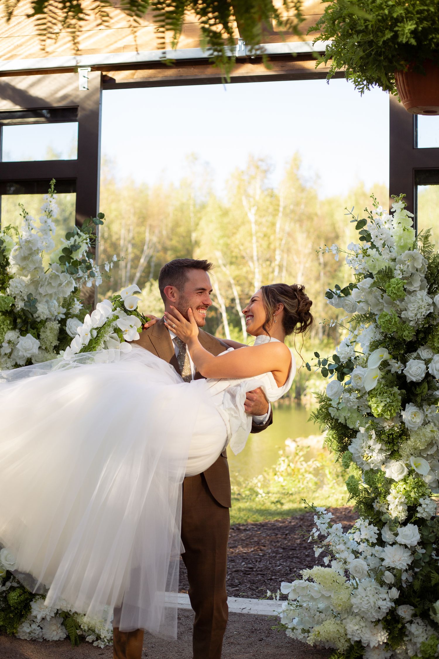 A romantic wedding moment captured under a floral arch with white flowers and greenery at sunset.