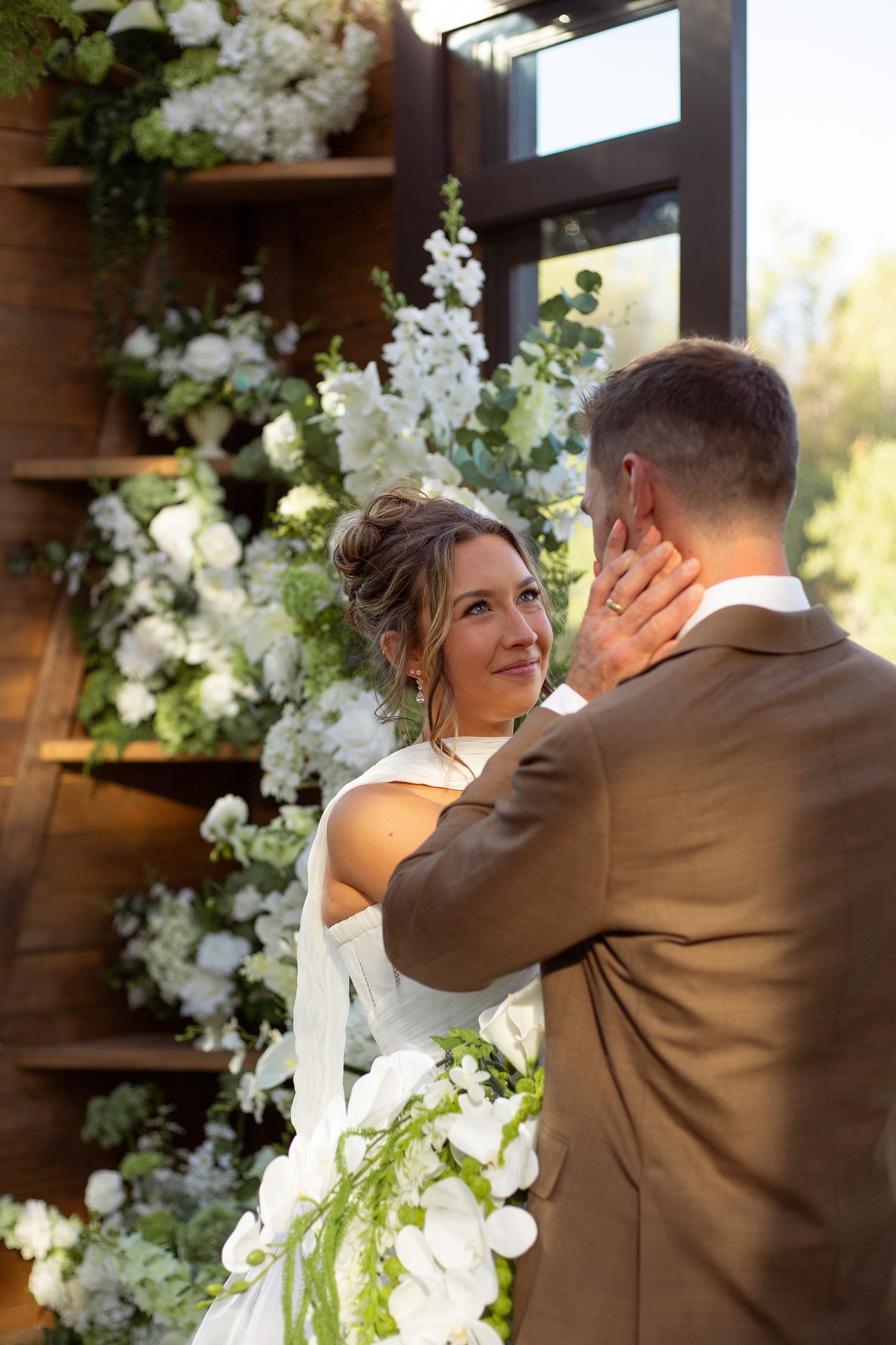A tender wedding moment captured in front of an elegant white floral arch with natural lighting.