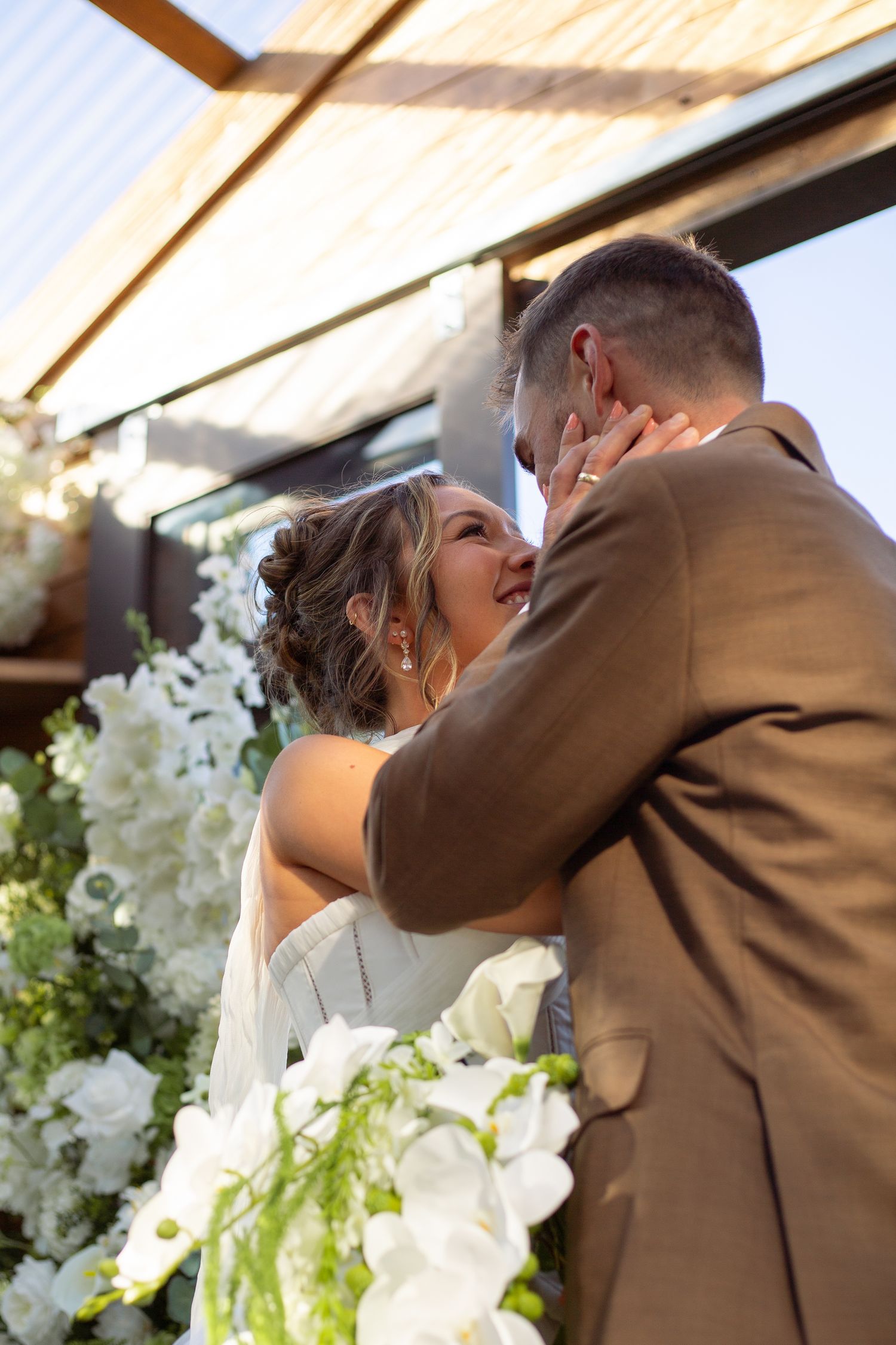A tender wedding moment captured under white floral arrangements and natural lighting.
