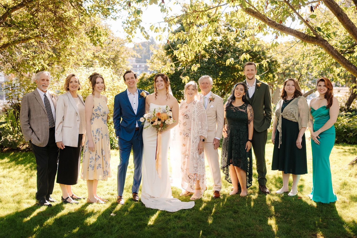 Wedding party poses together outdoors on a sunny day under trees with golden sunlight filtering through the leaves.