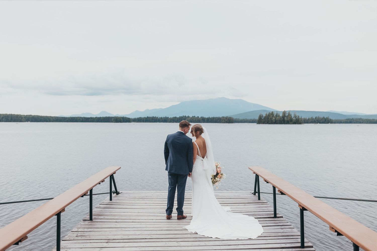 Long wooden pier stretches over calm lake water with mountains in the distance.