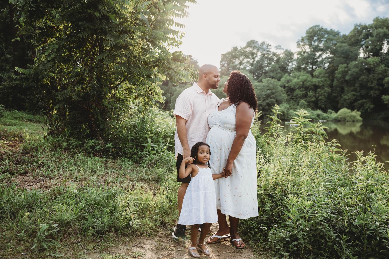 A family maternity photo with parents and young daughter in white outfits outdoors near a pond.