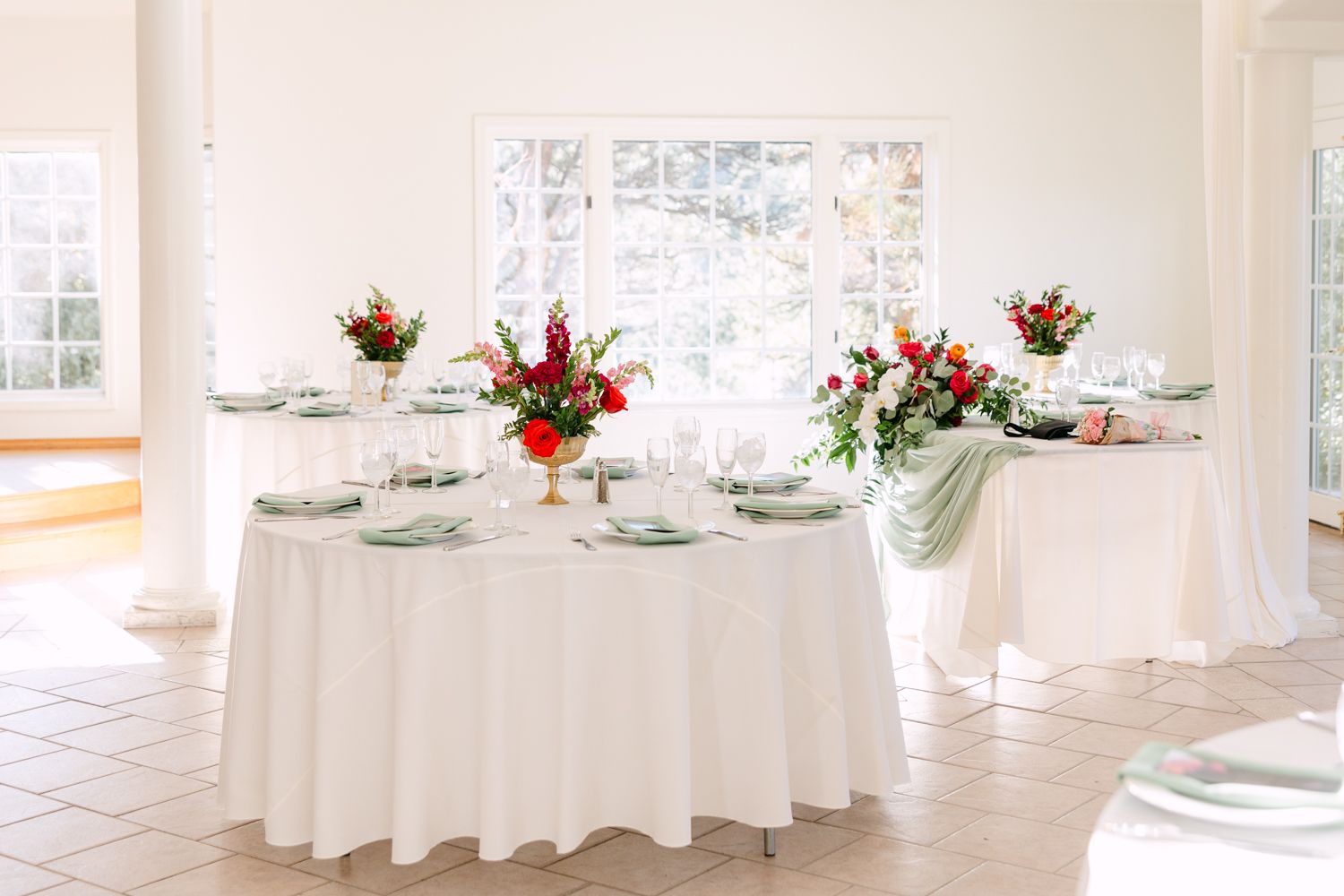 Round banquet tables with white tablecloths and floral centerpieces set up in an elegant white reception venue.