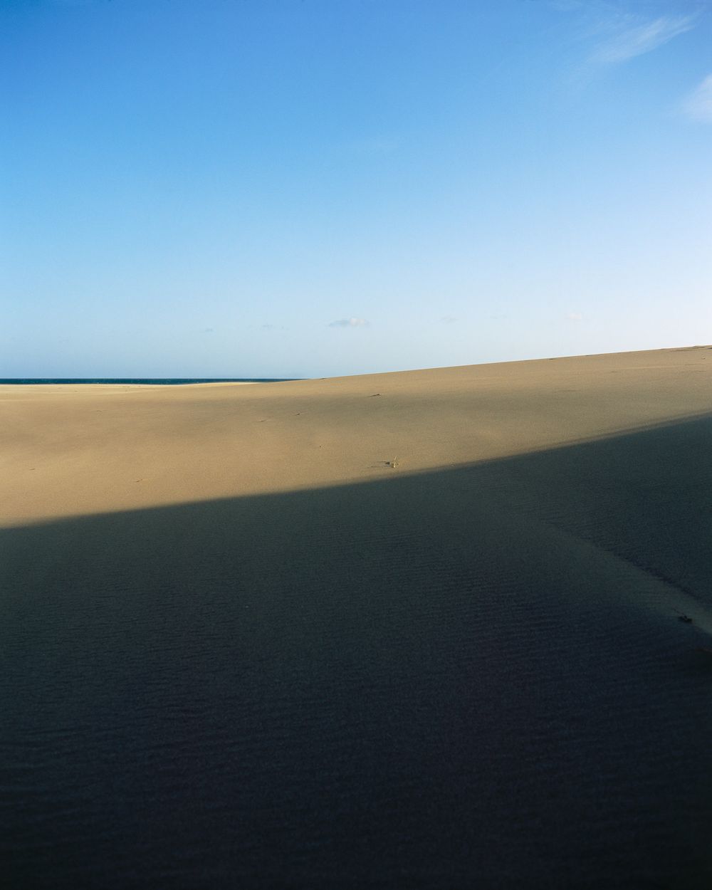 Desert landscape with rolling sand dunes casting dark shadows against a bright blue sky.