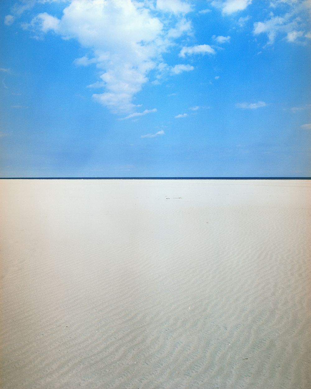 Rippled sand dunes stretch to the horizon under scattered white clouds and blue sky.
