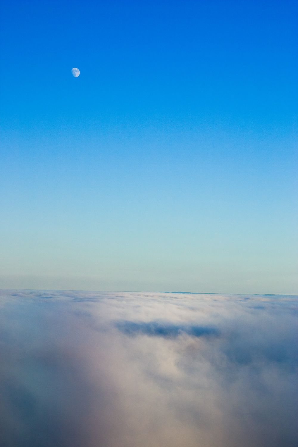 Moon visible in clear blue sky above a sea of white clouds stretching to the horizon.