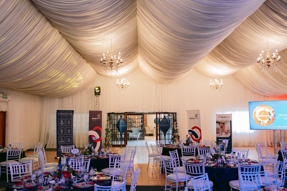 Elegant event venue with draped white ceiling, chandeliers, decorated round tables, and chairs set for a formal banquet.