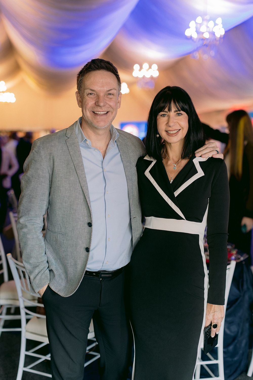 A smiling man in a gray blazer and woman in black wrap dress pose together at an elegant event venue.