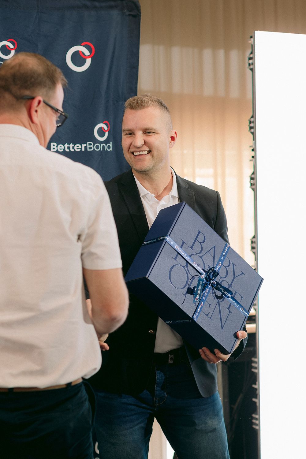 Man in blazer smiling while receiving a blue baby gift box at a BetterBond corporate event.