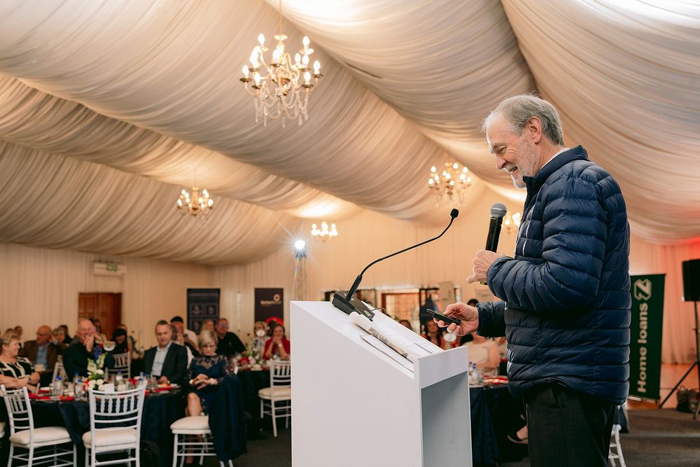 Man speaking at podium during formal banquet event in elegant draped tent venue with chandeliers.