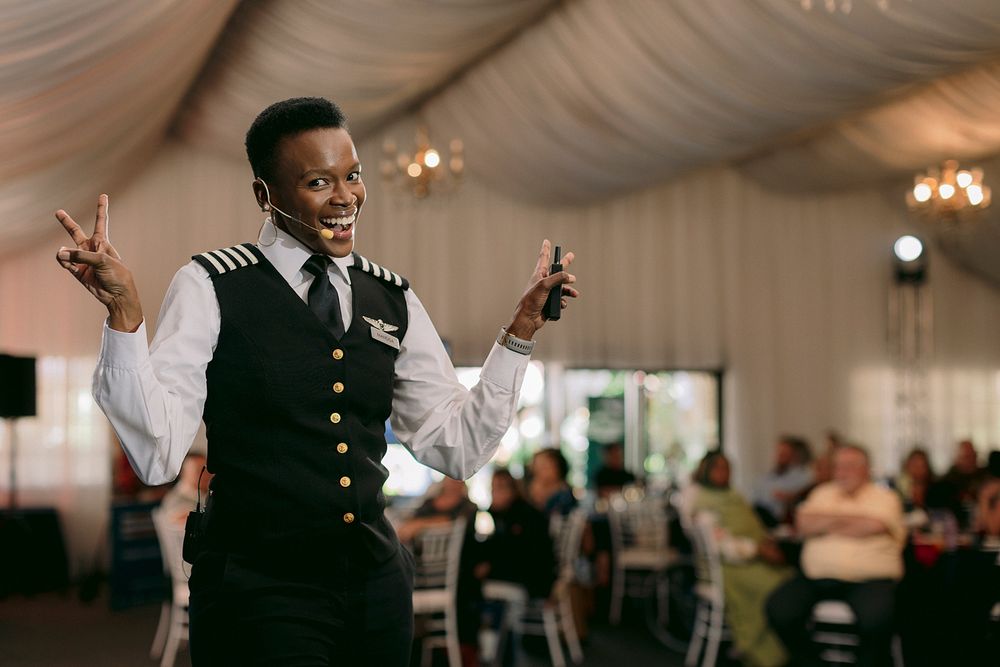 Smiling waiter in vest and uniform gestures at an elegant wedding reception venue with guests seated in background.
