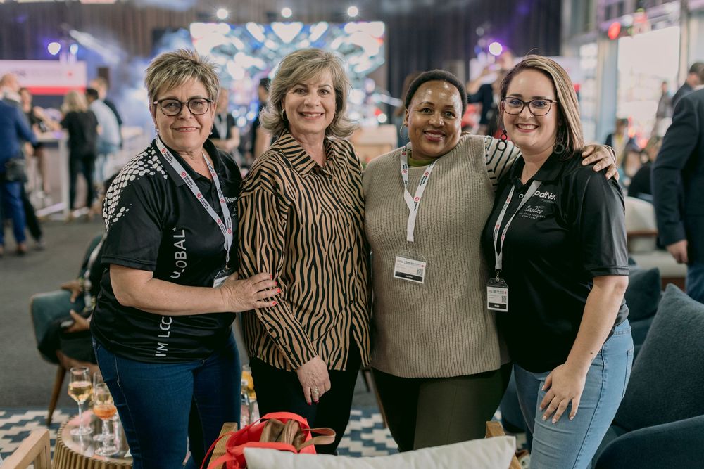 Four women smiling together at an indoor corporate networking event with colorful decor.