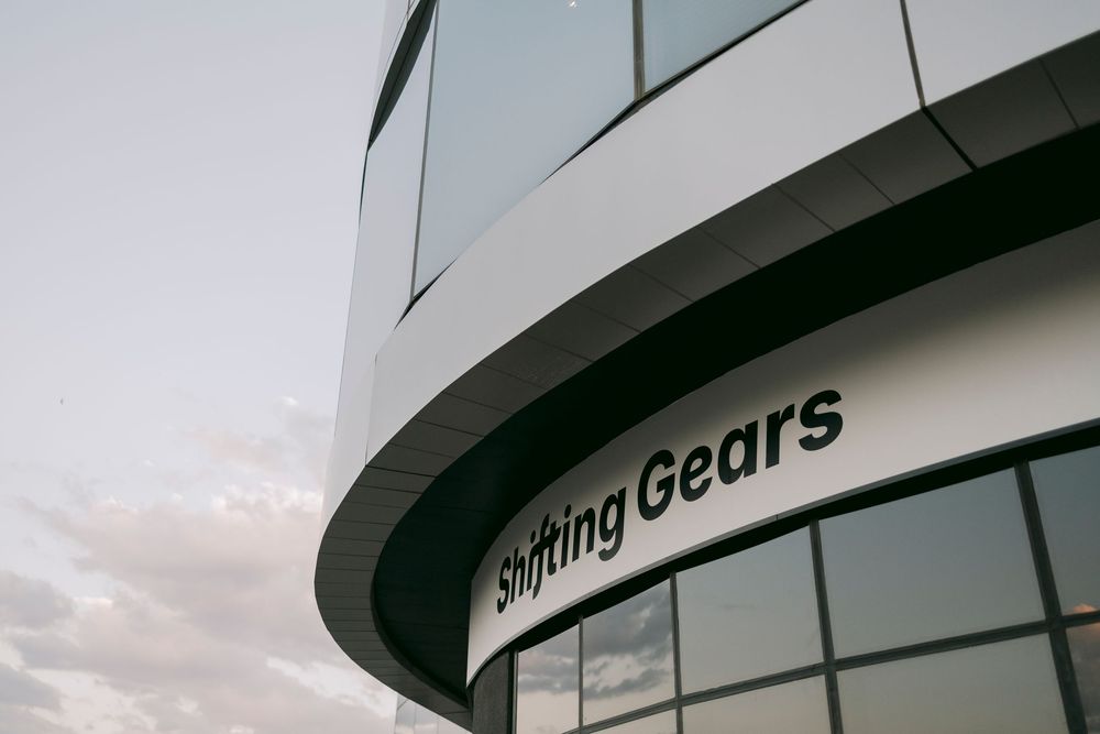 Close-up of a curved modern building facade with Shifting Gears signage against cloudy sky.