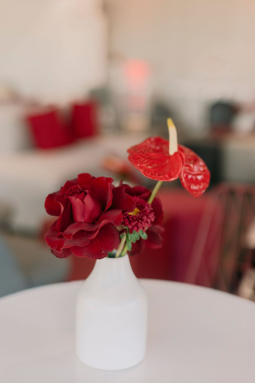Red anthurium and dark roses in a white vase on a white table in a softly blurred event venue.