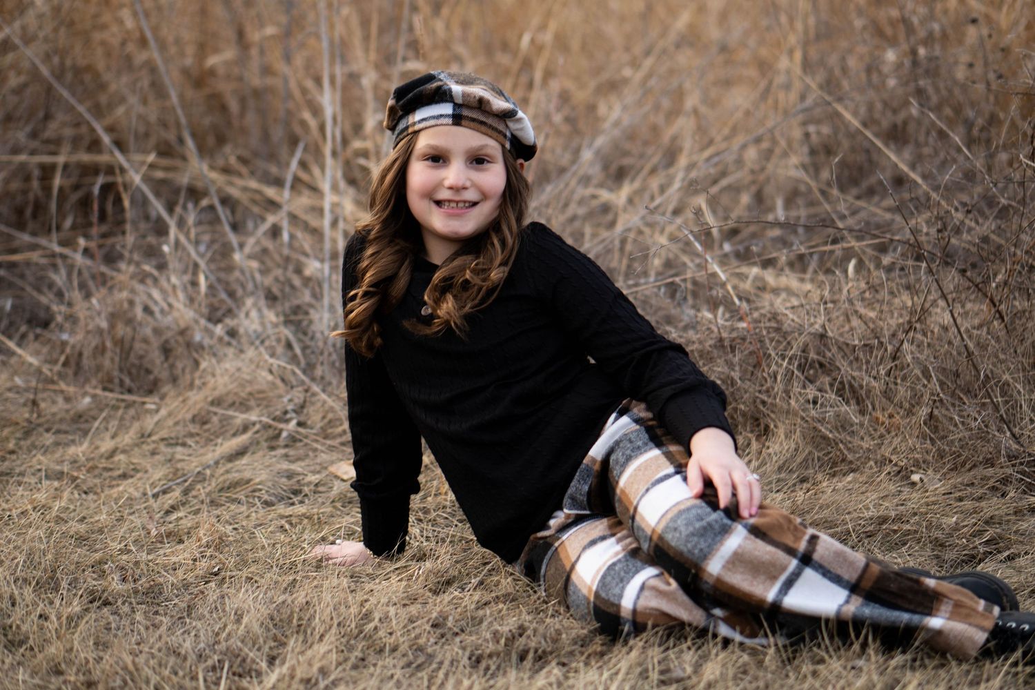 Young girl sitting in tall dry grass, wearing black top and plaid pants with a matching headband, smiling.