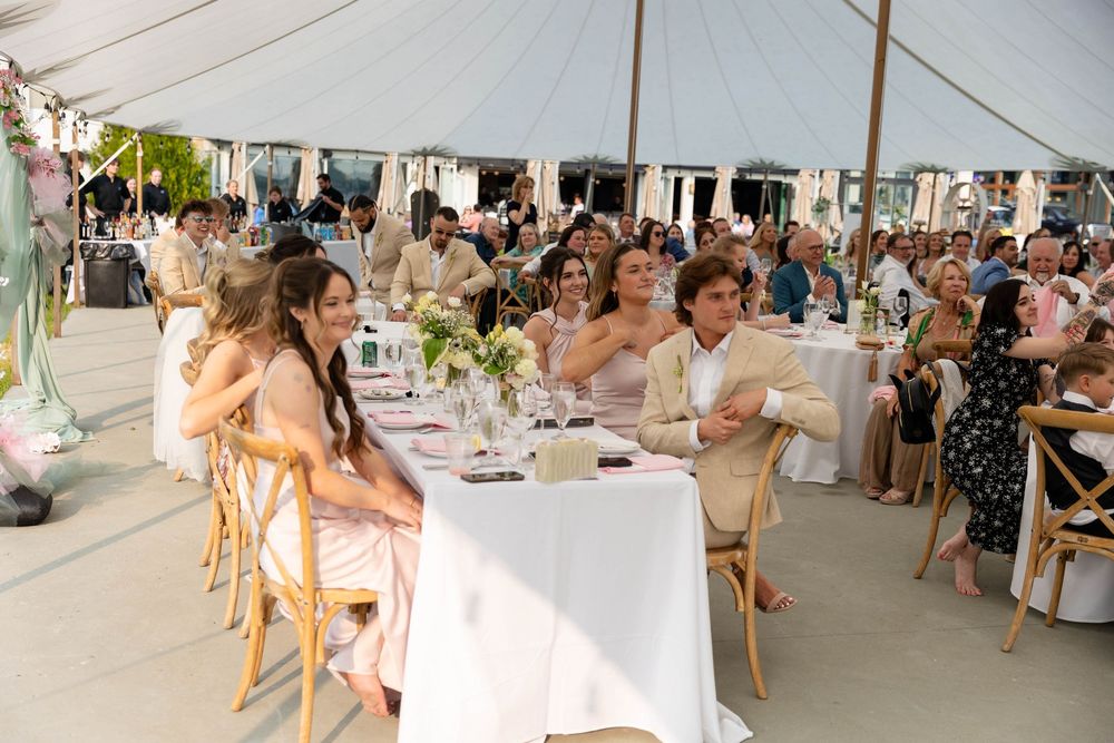 Wedding reception guests seated at elegant white-clothed tables under an outdoor tent with floral centerpieces.