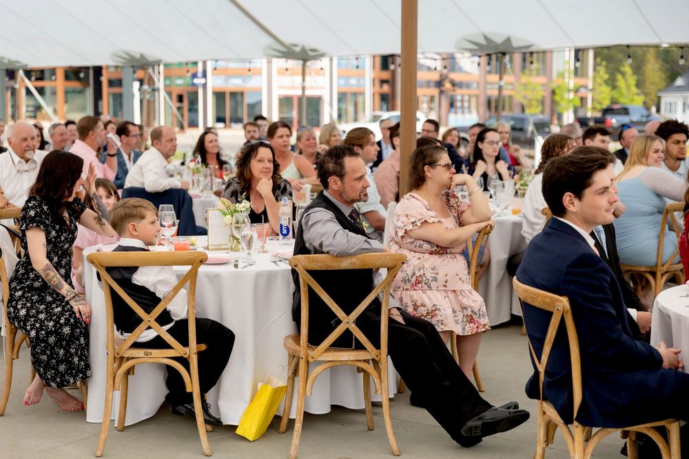 Wedding reception guests seated at long tables with cross-back chairs enjoy an outdoor celebration under string lights.