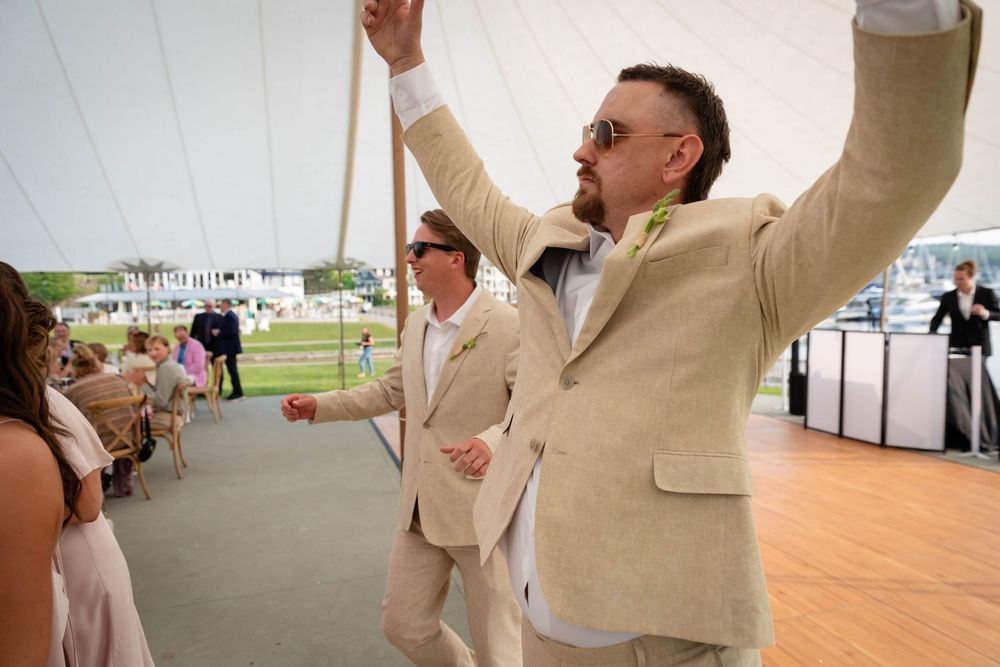 A group of people in beige suits dance and celebrate together under a white tent at an outdoor event.