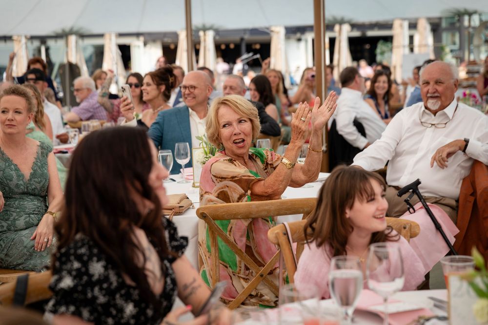 Wedding guests laugh and smile while seated at reception tables during a celebration event.