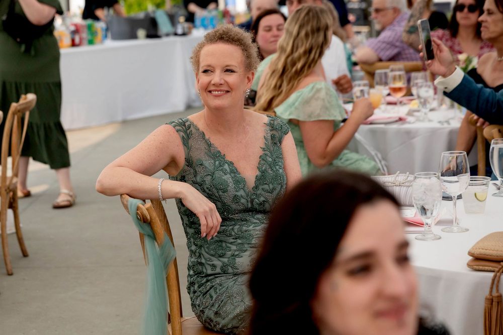Wedding guest in a sage green lace dress smiles while seated at an outdoor reception table.