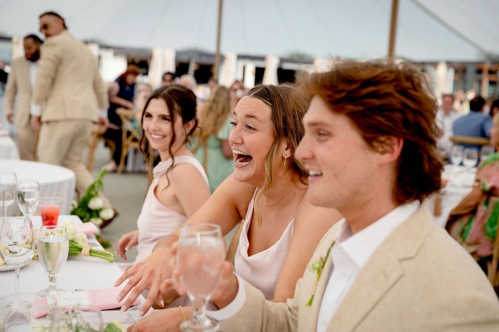 Wedding guests seated at a table under a tent share a joyful moment during the reception celebration.