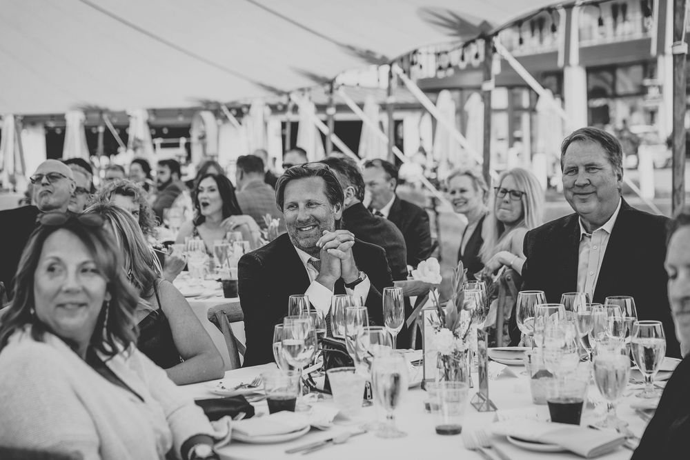 Wedding guests seated at long tables enjoy a reception dinner and conversation in an elegant white tent setting.