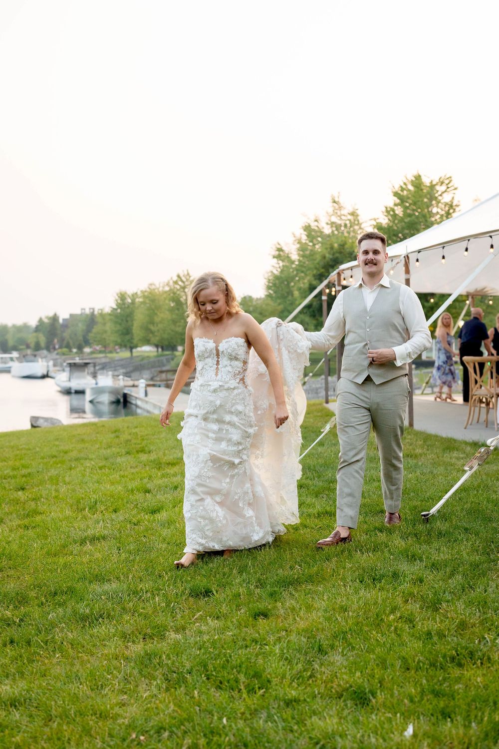 A couple in wedding attire playfully runs and poses on a grassy hillside near a waterfront on their special day.