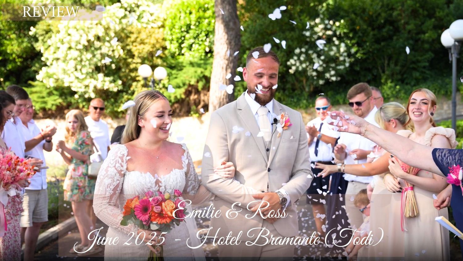 Wedding party celebrates with confetti toss as newlyweds walk down garden path with pink bouquet.