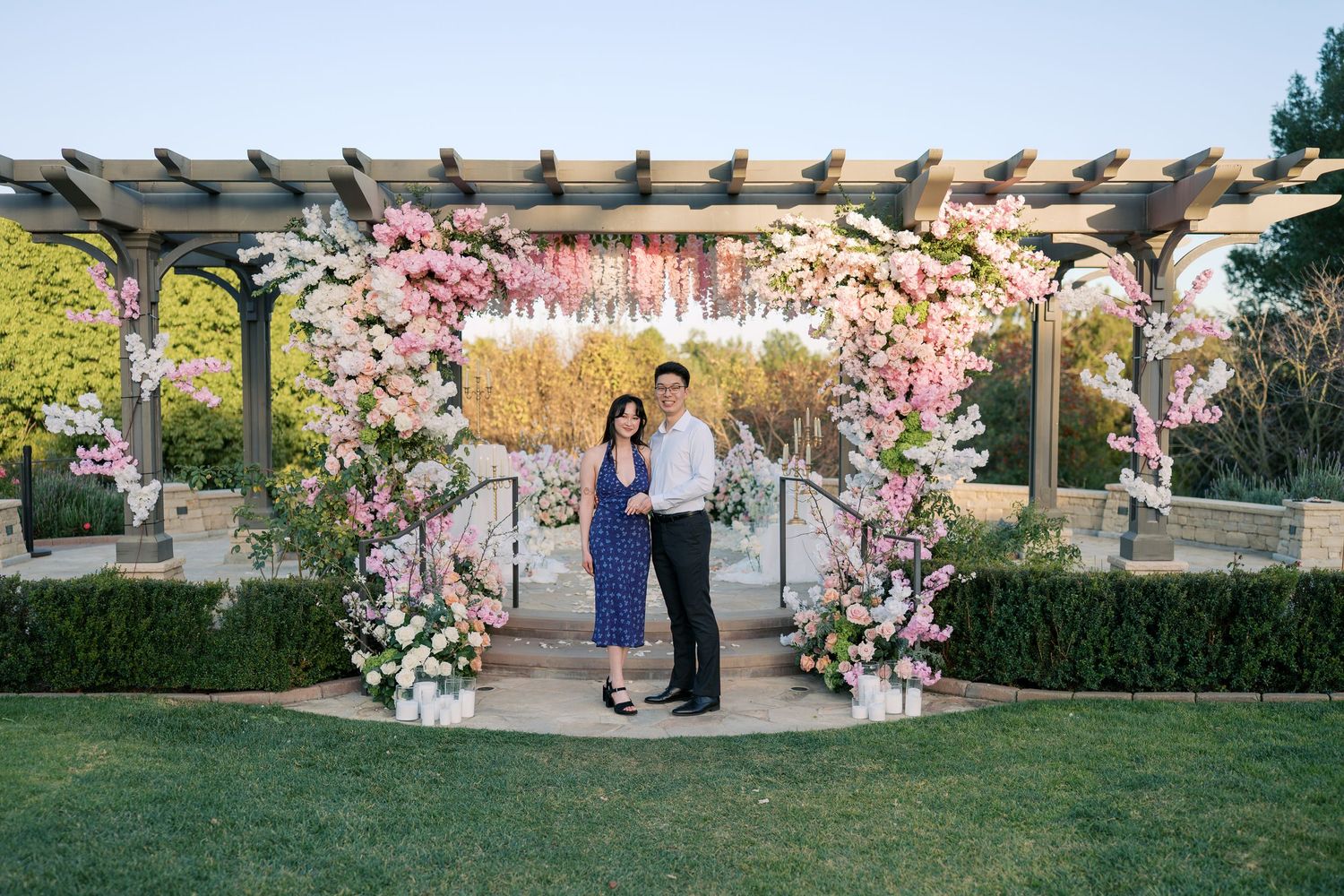 A romantic outdoor garden wedding ceremony setup with pink cherry blossom floral arrangements and a wooden pergola.