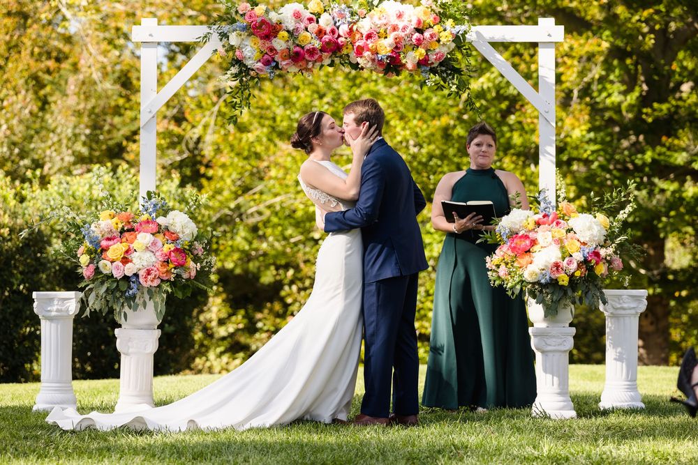 Wedding ceremony taking place outdoors under a white arbor with floral arrangements and greenery.
