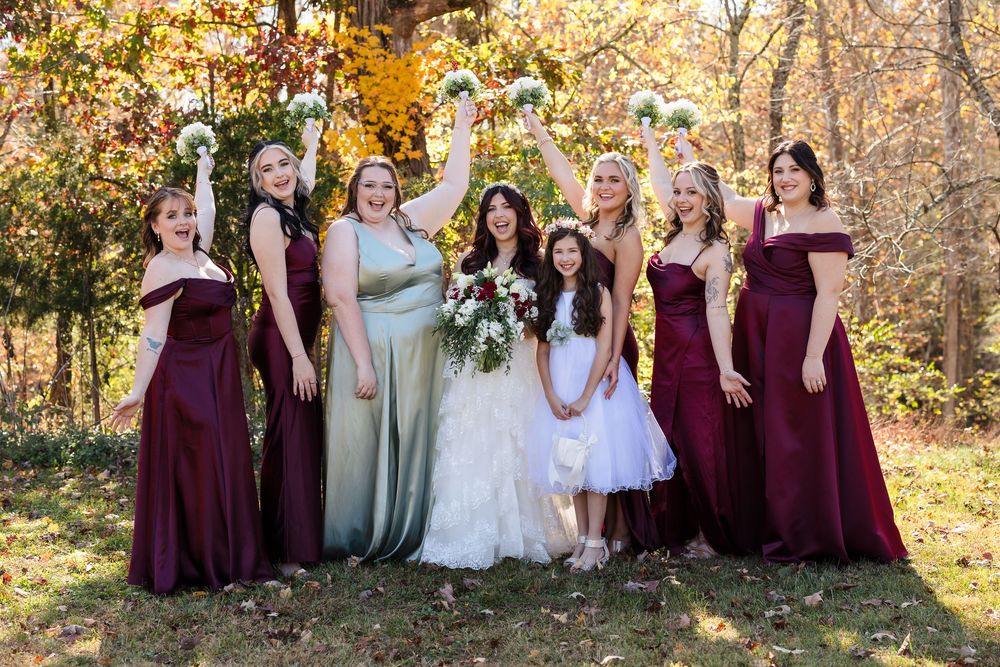 Large bridal party in burgundy and sage colored dresses poses together against fall foliage backdrop.