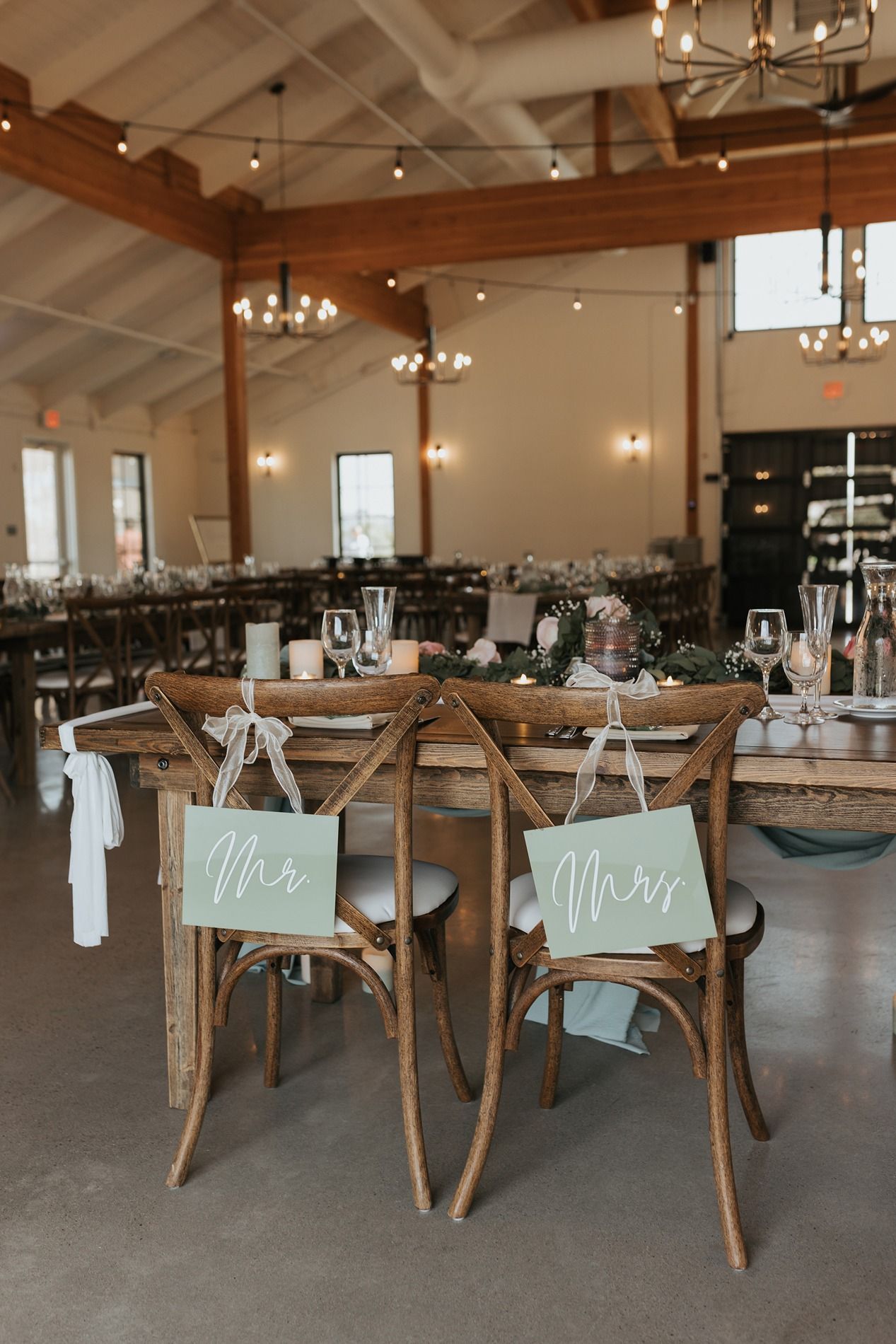 Rustic wooden cross-back chairs with 'Mr' and 'Mrs' signs arranged in an elegant wedding reception venue with chandeliers at Canopy Grove.