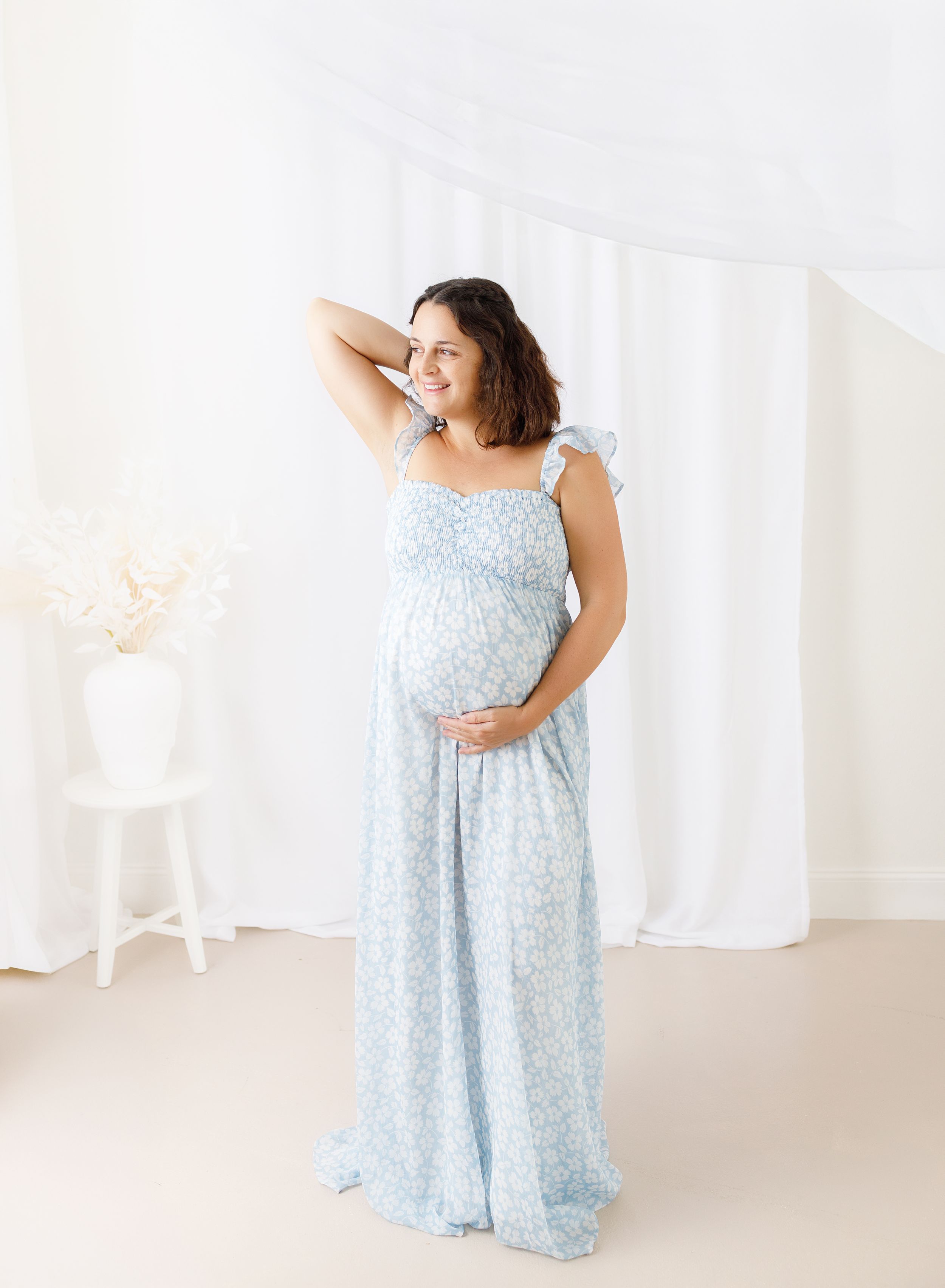 A pregnant woman in a light blue floral maxi dress poses gracefully against a white backdrop in a photo studio setting.