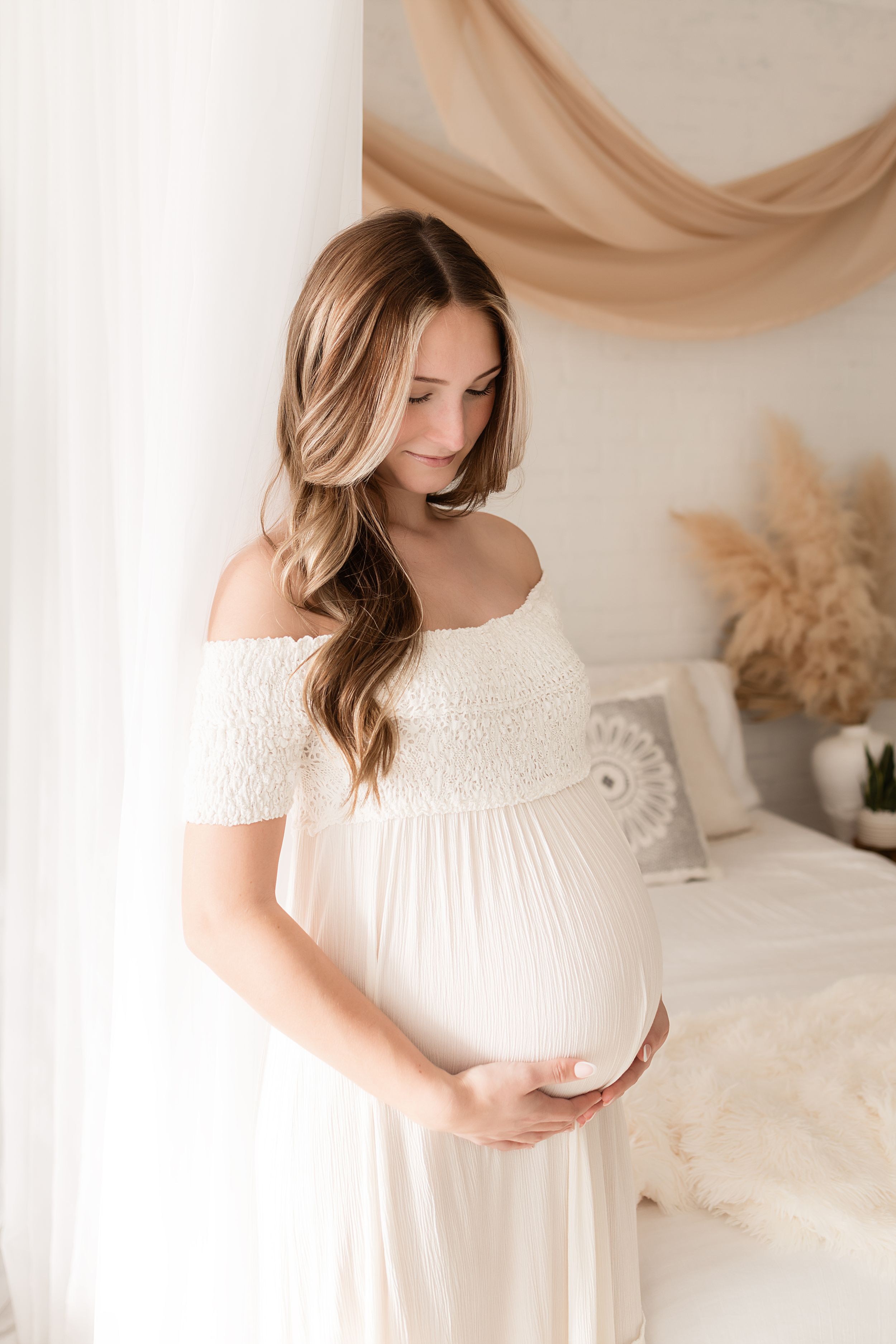 Expecting mother in flowing white dress poses softly in bright studio with draped fabric and pampas grass decor.