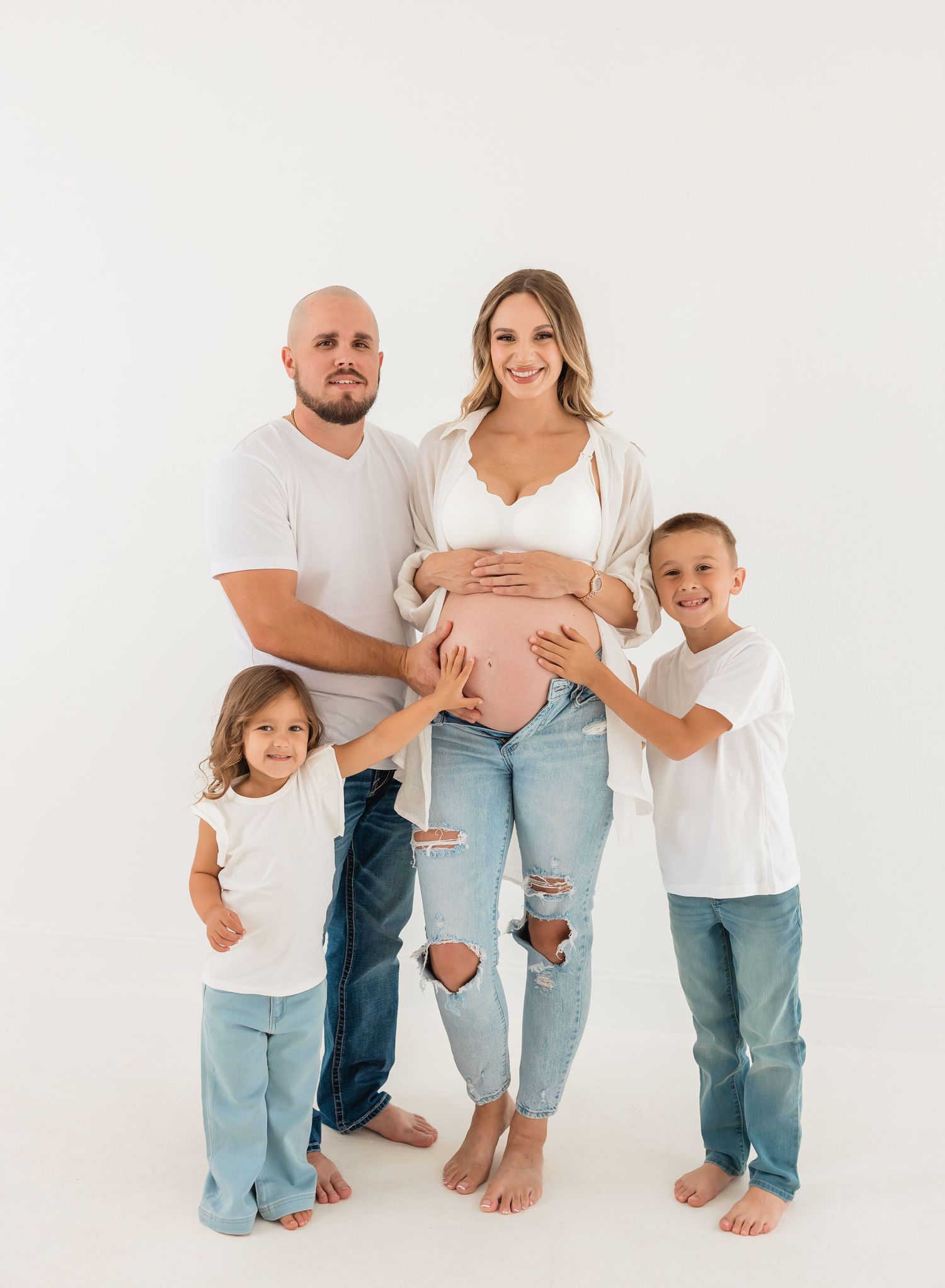 A family of four poses together against a white background, with the expectant mother wearing ripped jeans and a cardigan.