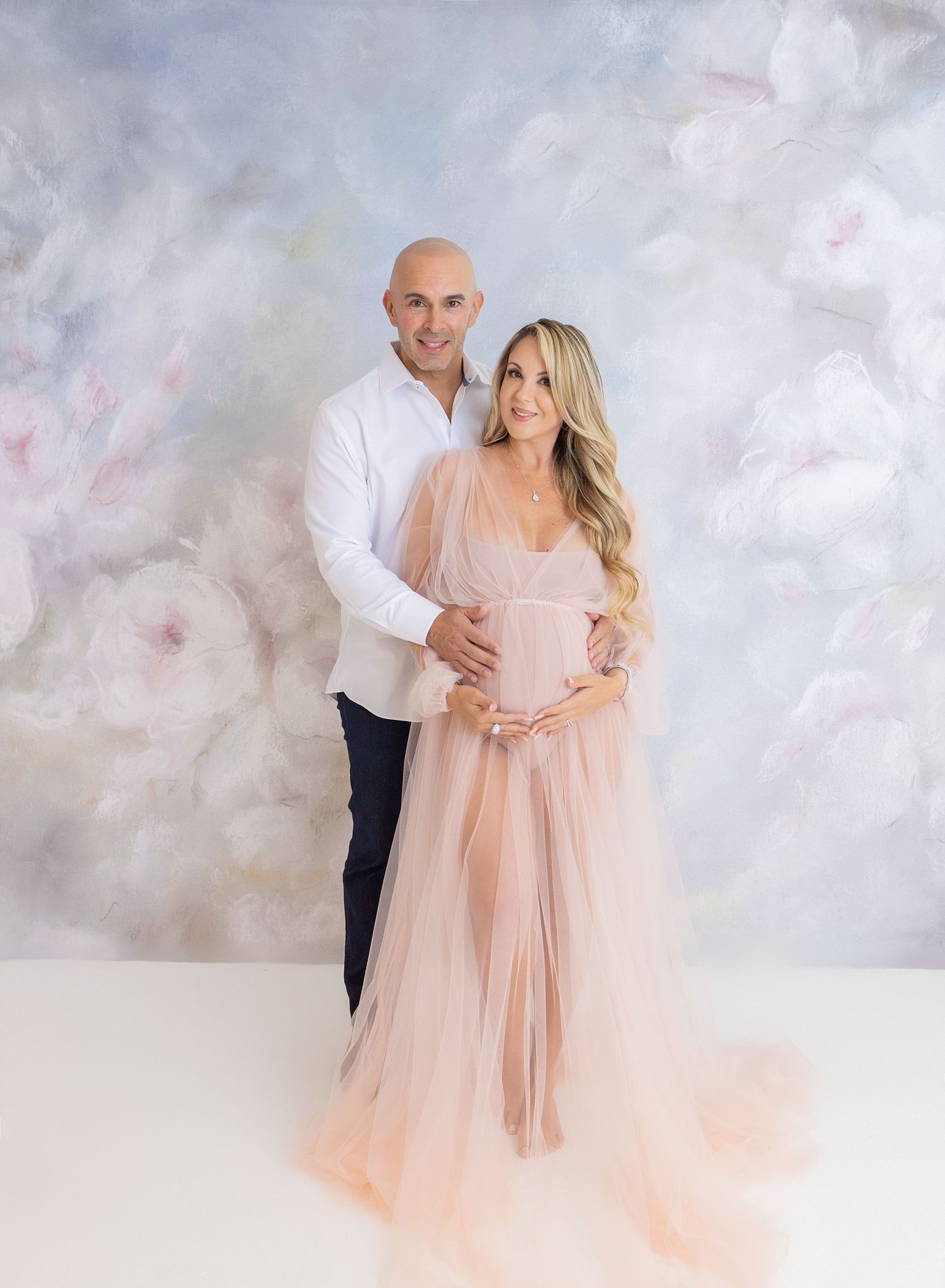 A couple embraces in a dreamy setting with soft pink tulle dress and white backdrop creating an ethereal maternity photo.