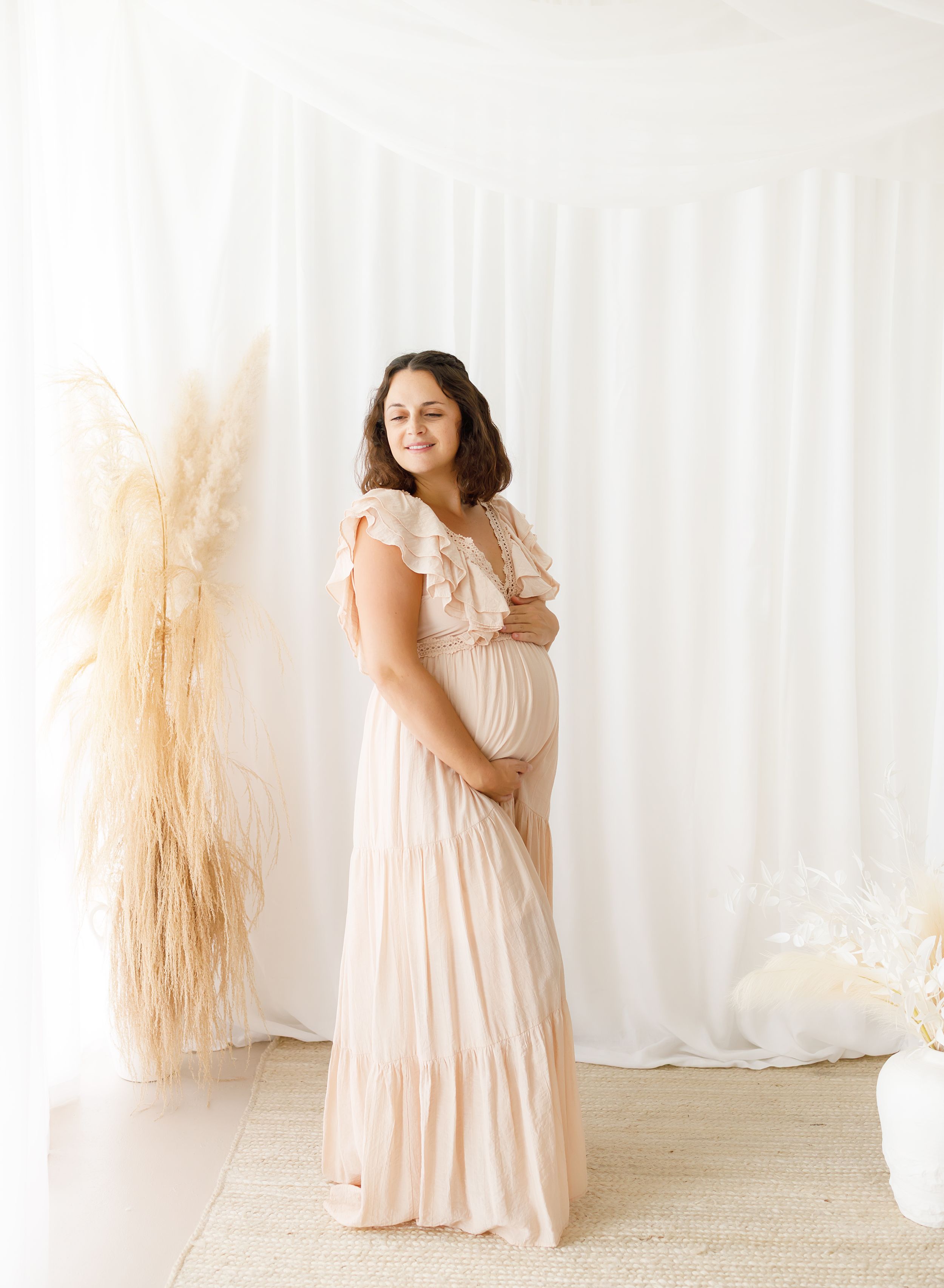 Woman in a flowing blush pink tiered dress poses gracefully against a white backdrop with dried pampas grass decor.