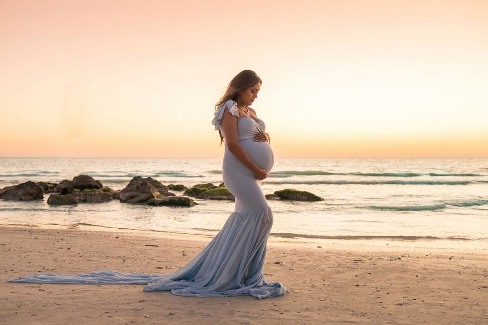 Pregnant woman in flowing gray gown poses on beach at golden sunset, cradling baby bump near ocean waves.
